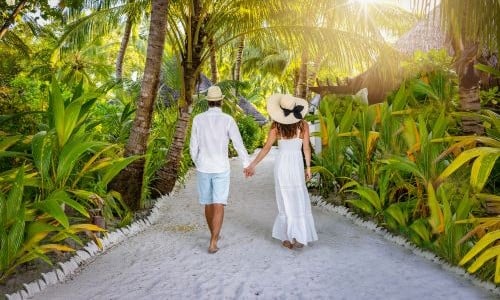 A couple holding hands walking, surrounded by white sand and palm trees