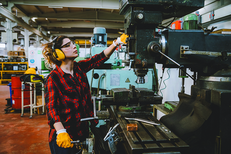 A person wearing yellow gloves and headphones and safety glasses standing next to a machine