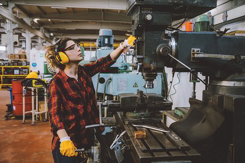 A person wearing yellow gloves and headphones and safety glasses standing next to a machine
