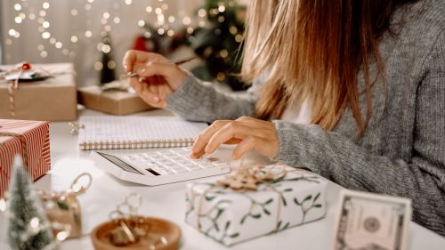 Woman using a calculator to figure out holiday expenses surrounded by Christmas presents