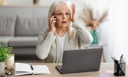 Surprised shocked mature woman talking on mobile phone in front of a laptop